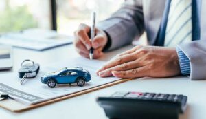 A person holding auto loan documents and car keys next to a parked modern car.
